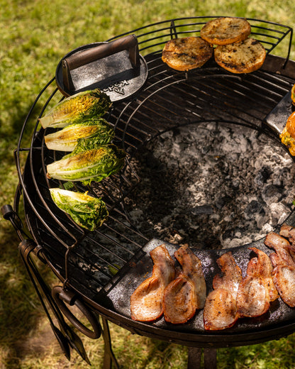 Grill with various foods being cooked, including vegetables and meats, with lettuce warming on a grassy background.