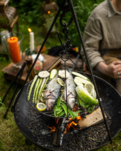 Hanging grill grate over fire pit with fish, greens, and sliced vegetables being grilled outdoors