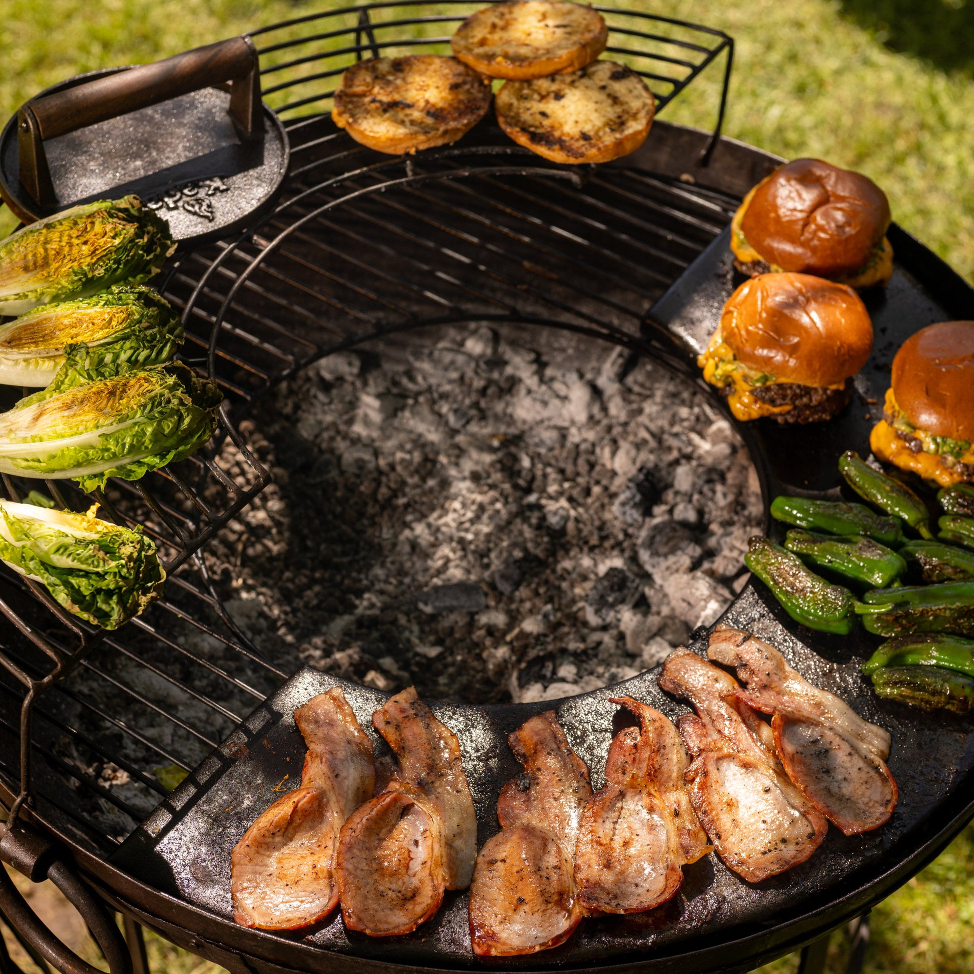 Barbecue grill with various foods including burgers, bacon, and vegetables on a natural outdoor setting.