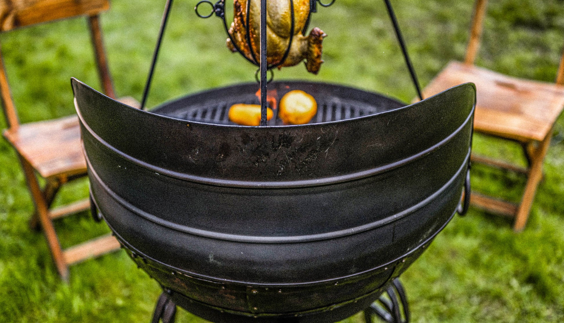 Kadai fire pit with curved wind guard panel protecting grill as chicken cooks over fire