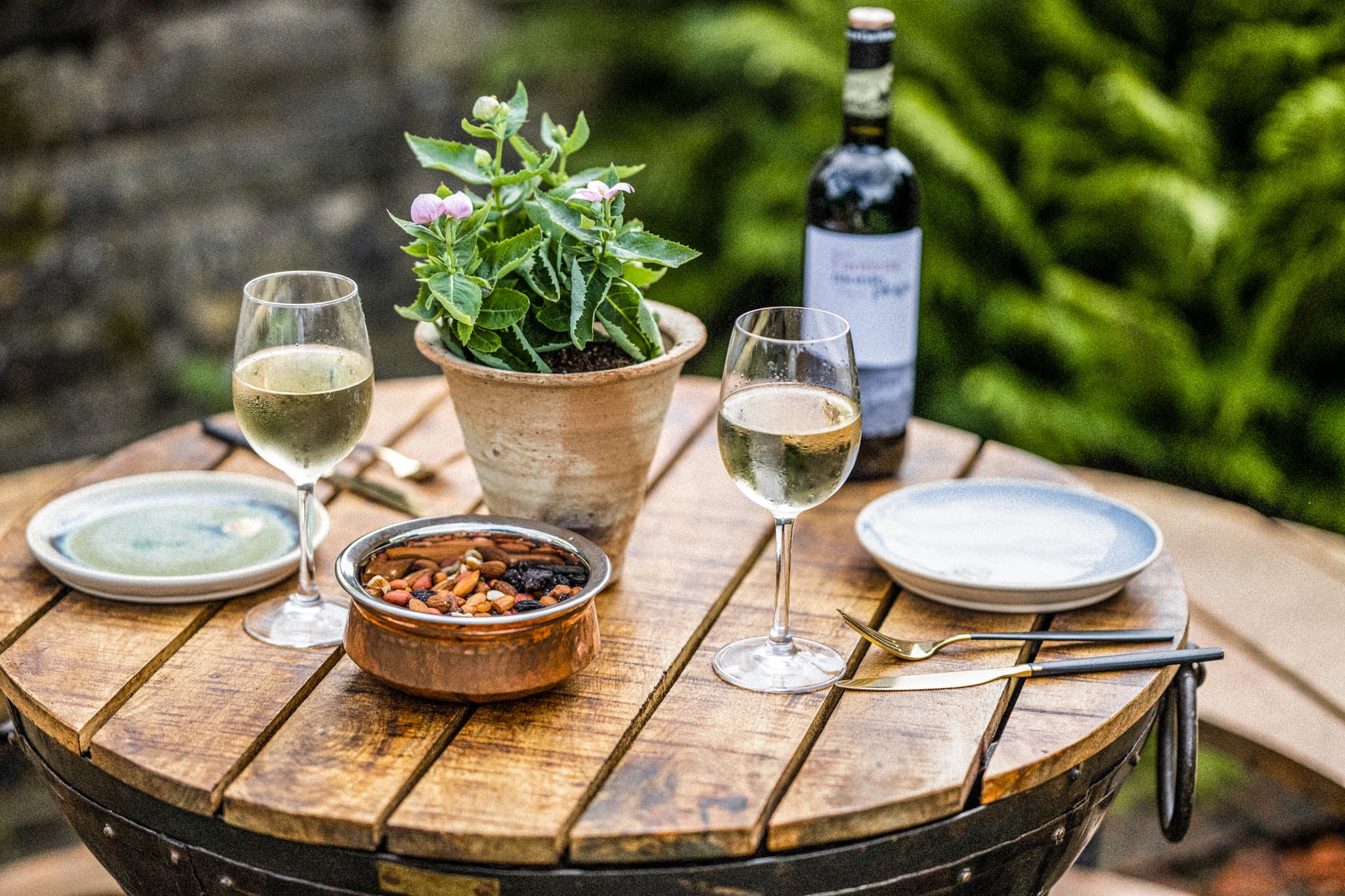 Round wooden Kadai table top set with wine glasses, snack bowl, plates, and a potted plant outdoors