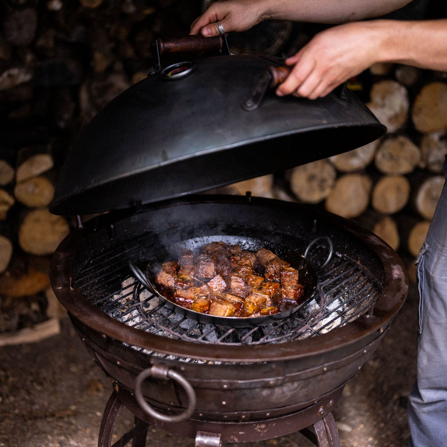 Opening a charcoal grill smoker to check slow-cooked meat inside, with steam rising from the pan