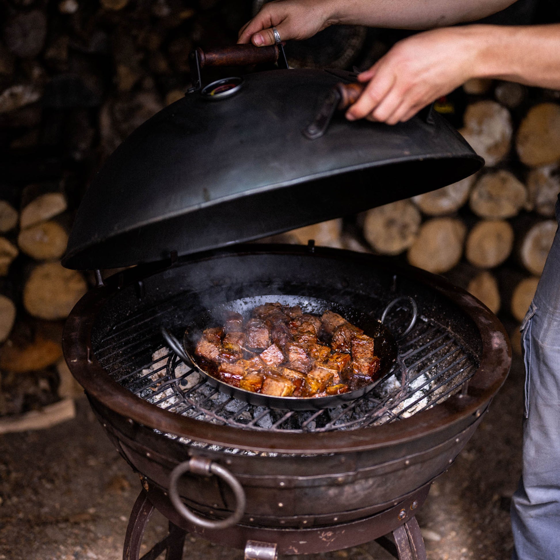 Opening a charcoal grill smoker to check slow-cooked meat inside, with steam rising from the pan