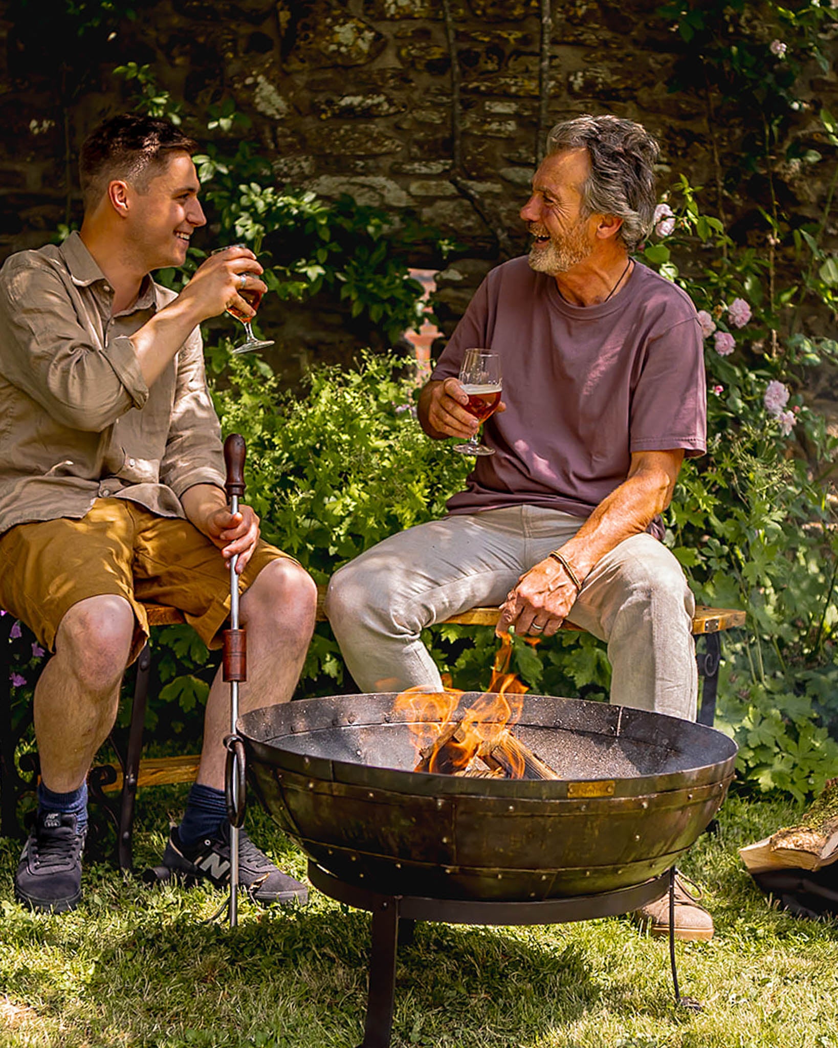 Medium sized charcoal BBQ fire bowl with open flames as two people sit beside it enjoying drinks in garden