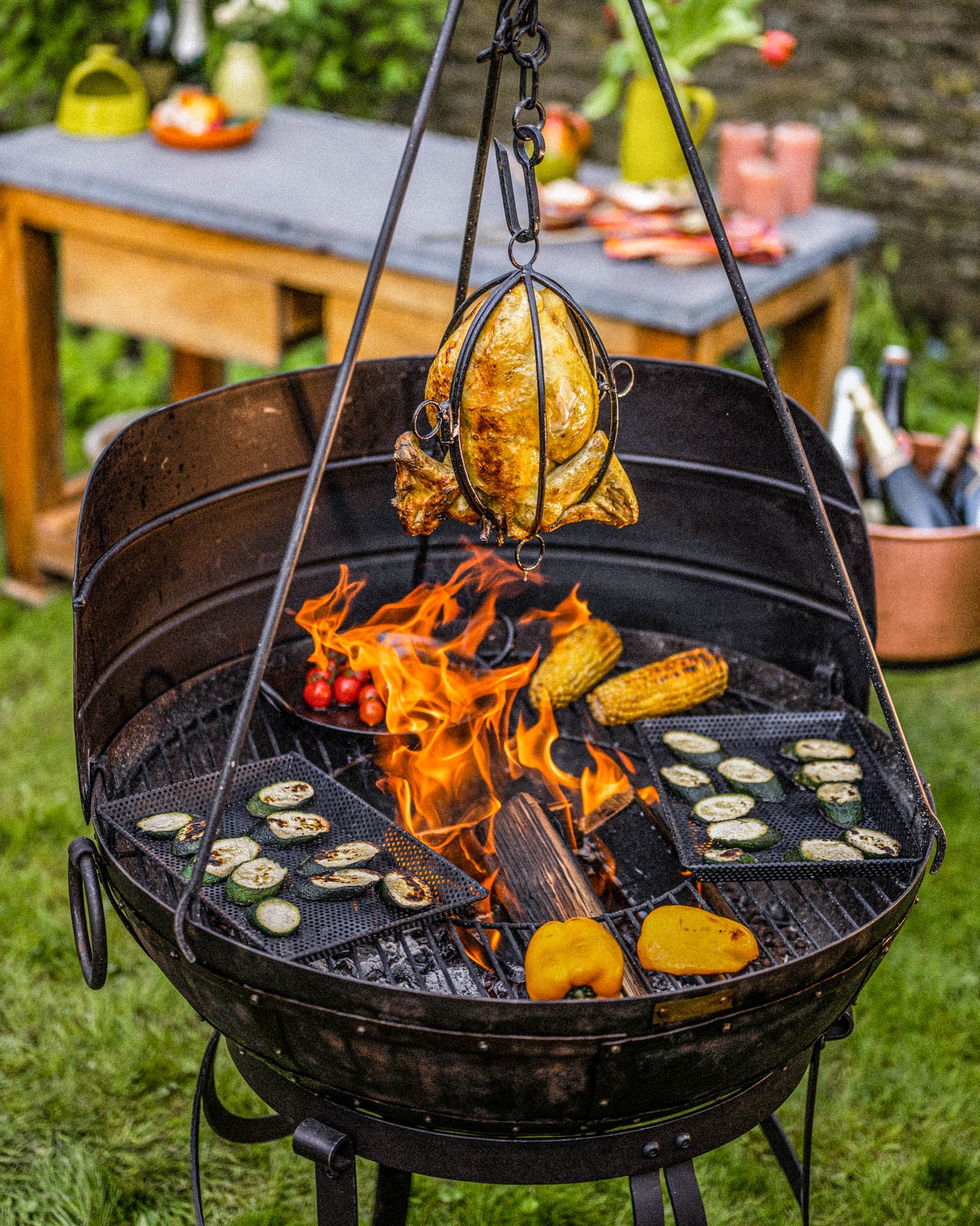 Kadai fire pit with wind guard and tripod cooking chicken over open flames with vegetables on grill