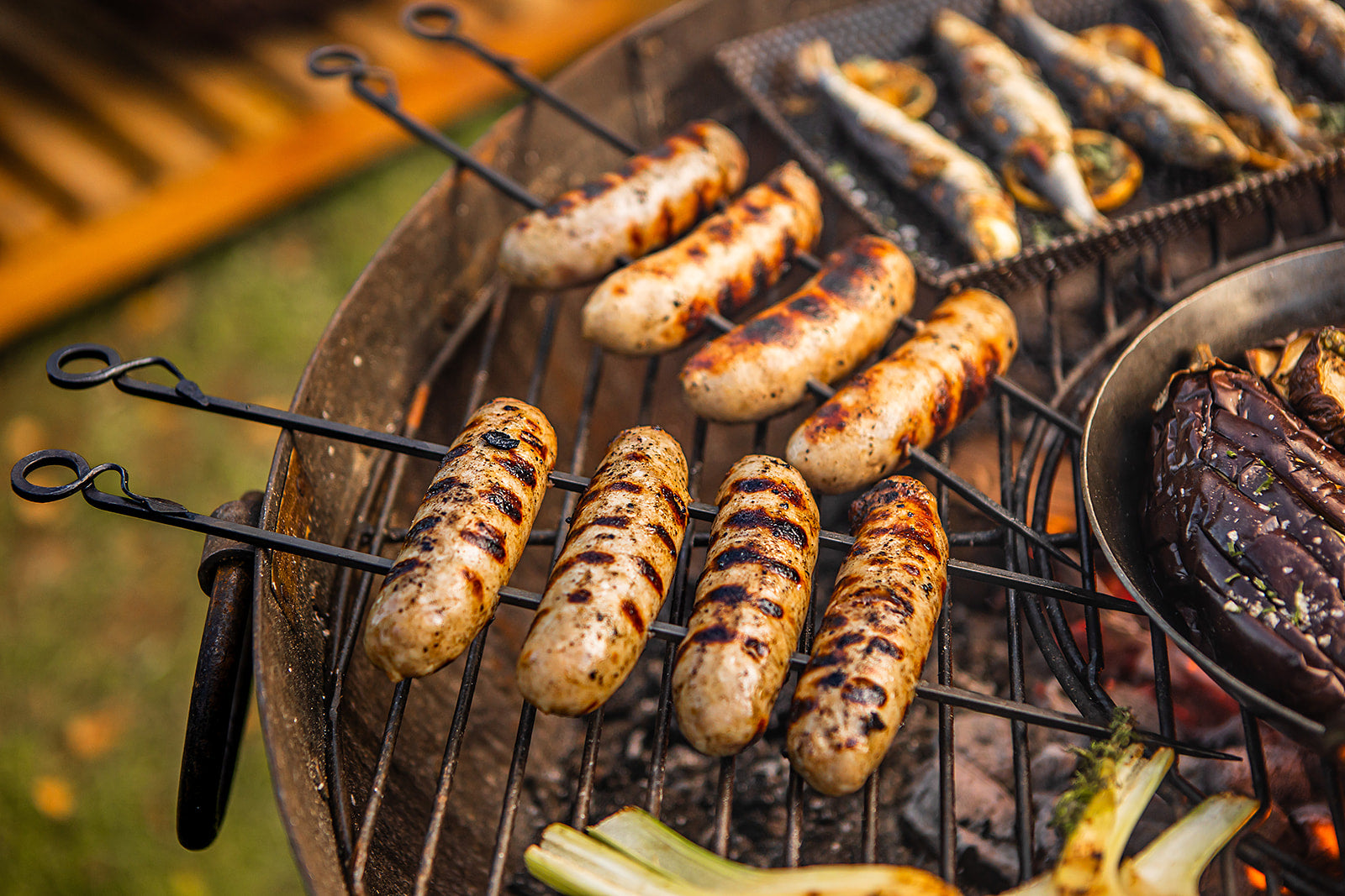 Grilled sausages cooking on metal BBQ skewers and rack over open coals in a rustic outdoor barbecue setup