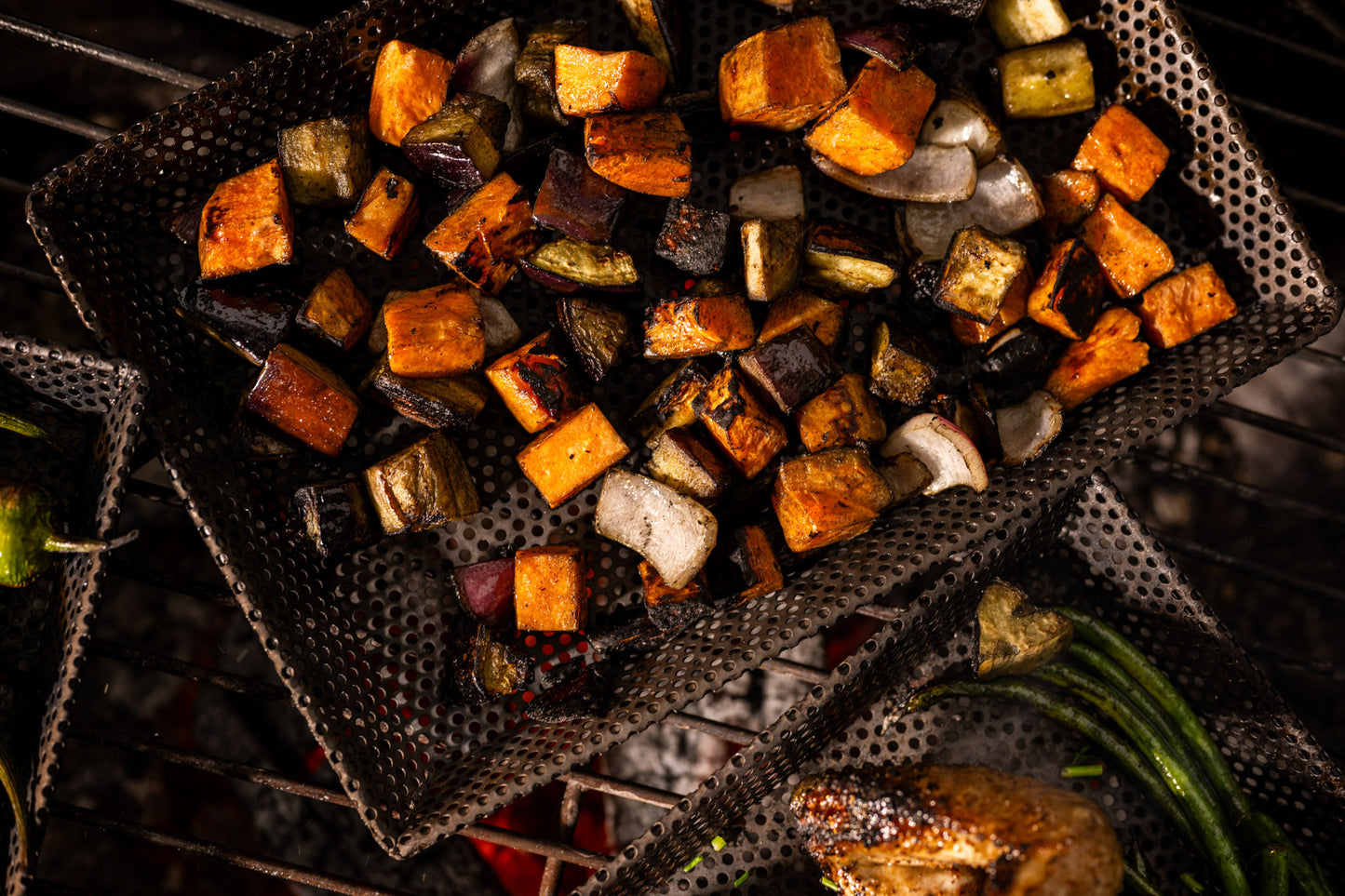 Diced vegetables roasting on perforated BBQ grill trays over glowing coals, including sweet potato, eggplant, onion, and zucchini with visible char marks and smoke.