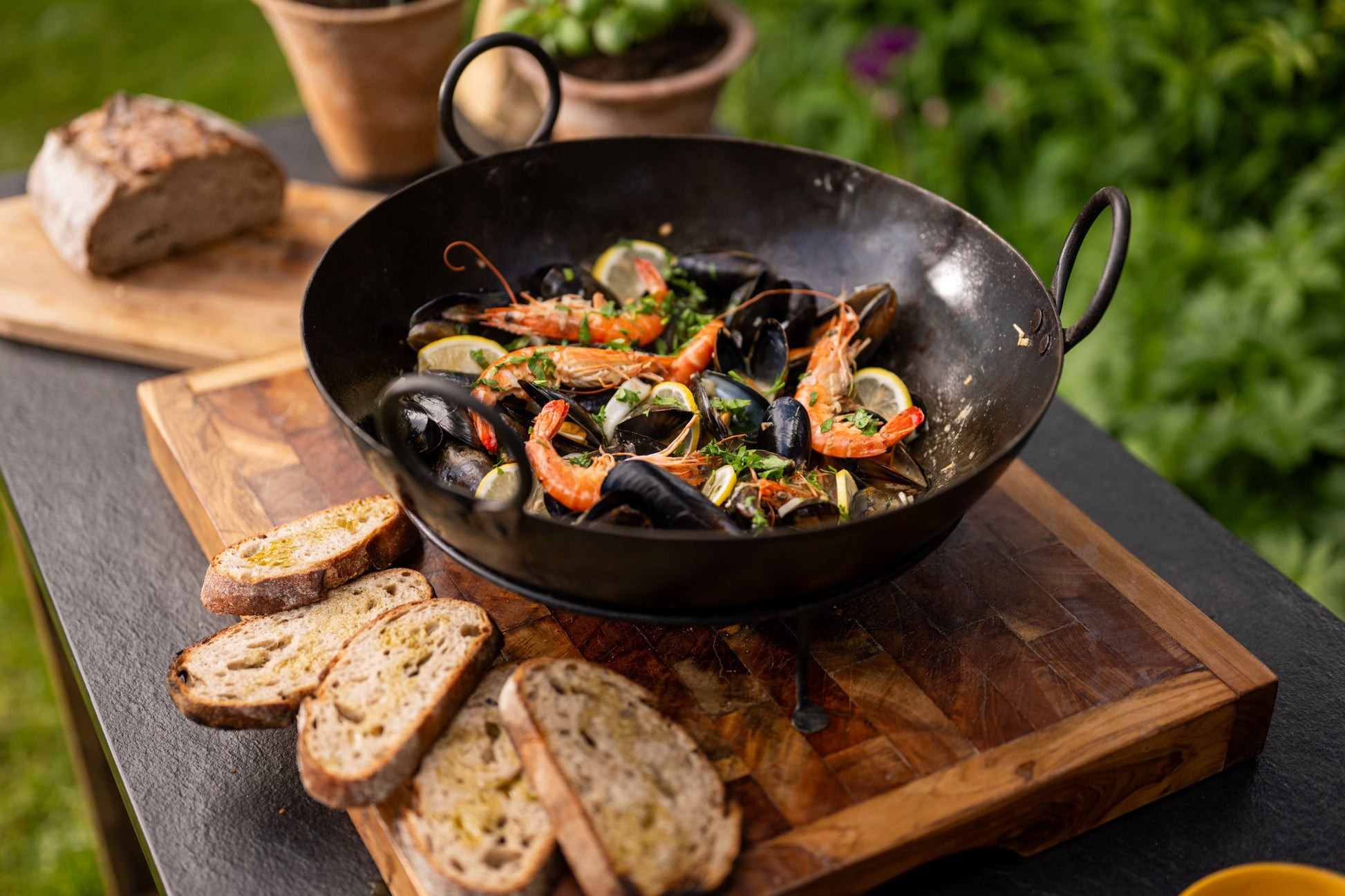 Seafood dish in a bowl on a wooden board with bread slices, outdoors.