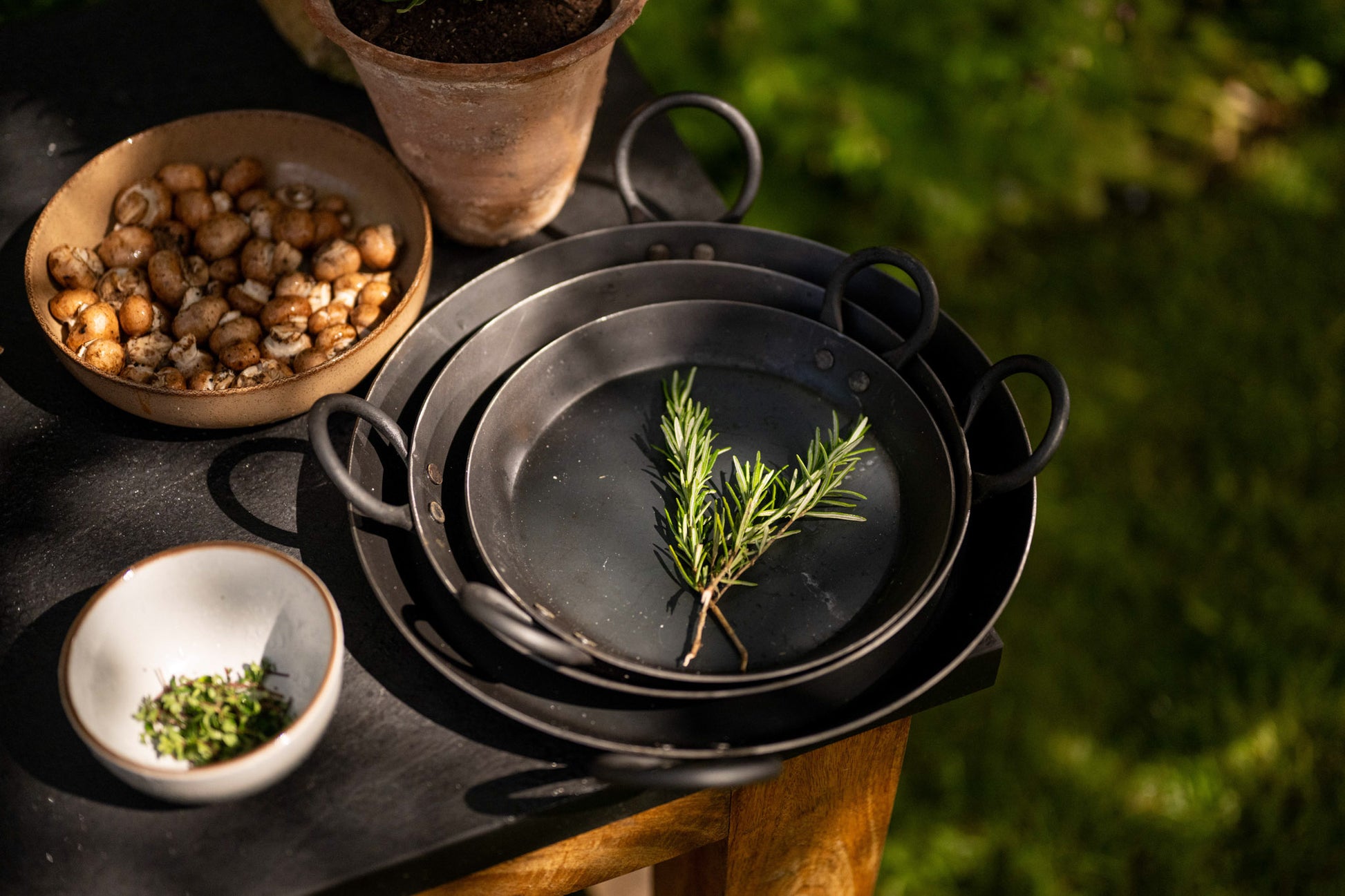 Stacked BBQ skillets prepared for outdoor cooking with fresh rosemary, mushrooms, and herbs on a rustic table.