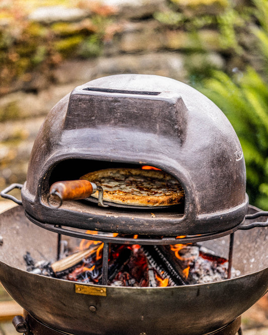 Outdoor pizza oven with a pizza inside, set over a fire pit with burning logs.