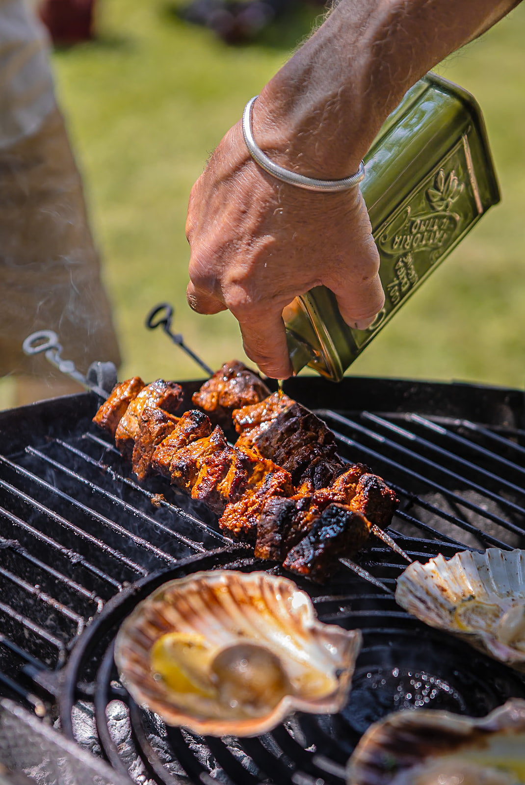Marinated meat skewers grilling over open flames while oil is drizzled onto the barbecue in an outdoor cooking scene