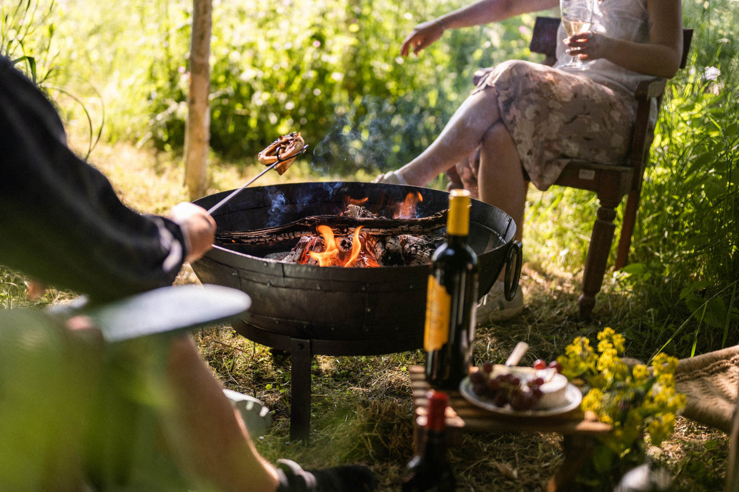 Person using a long toasting fork to warm food over an outdoor fire pit, while others relax nearby with drinks and snacks.