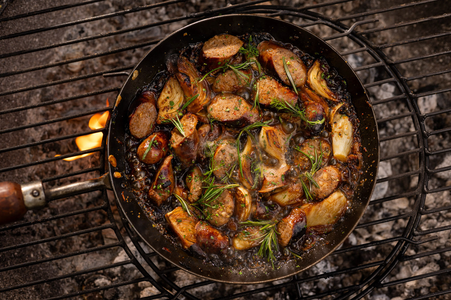 BBQ frying pan filled with sausages, caramelised onions and rosemary cooking over open flames on a Kadai firebowl barbecue grill