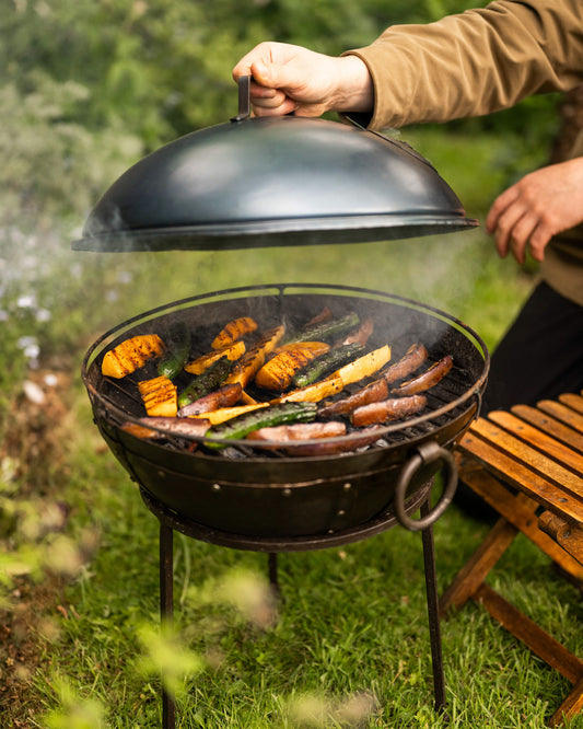 Person grilling vegetables and sausages on a barbecue grill outdoors.