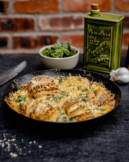 Close-up of a cheesy baked potato dish in a skillet, topped with herbs, with olive oil and garlic in the background