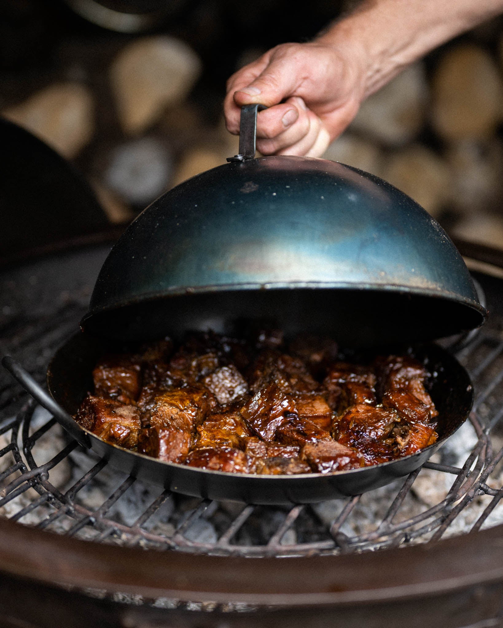 BBQ cloche being lifted to reveal sizzling smoked meat cooking on a grill over hot coals with rising steam