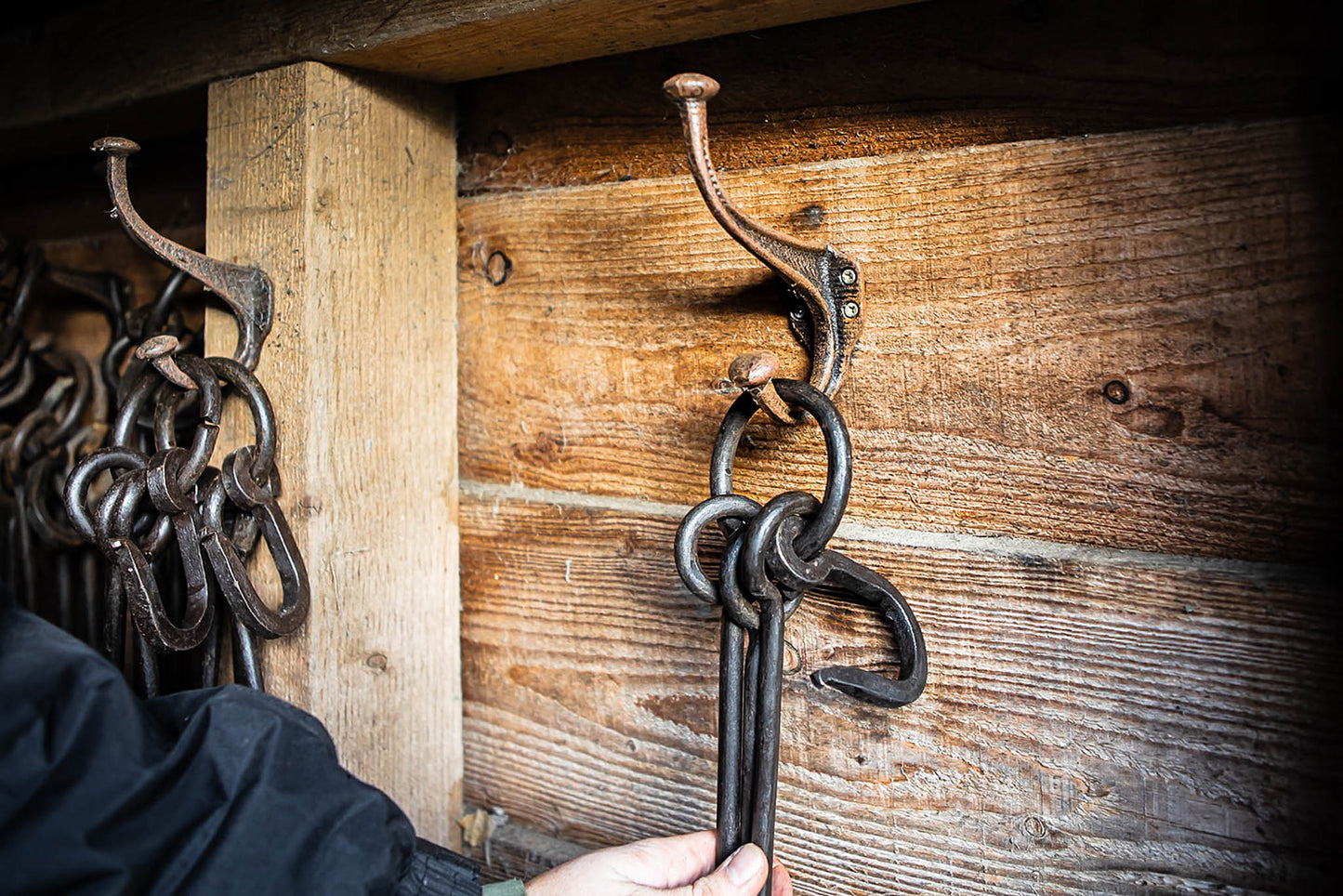 Hand placing black metal fire tools onto rustic cast iron wall hooks mounted on weathered wooden boards.