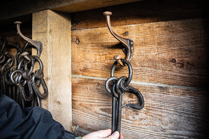 Hand placing black metal fire tools onto rustic cast iron wall hooks mounted on weathered wooden boards.