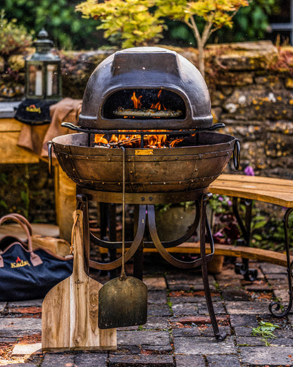 Wood fired oven set on a Kadai grill with flames burning underneath, surrounded by outdoor cooking tools