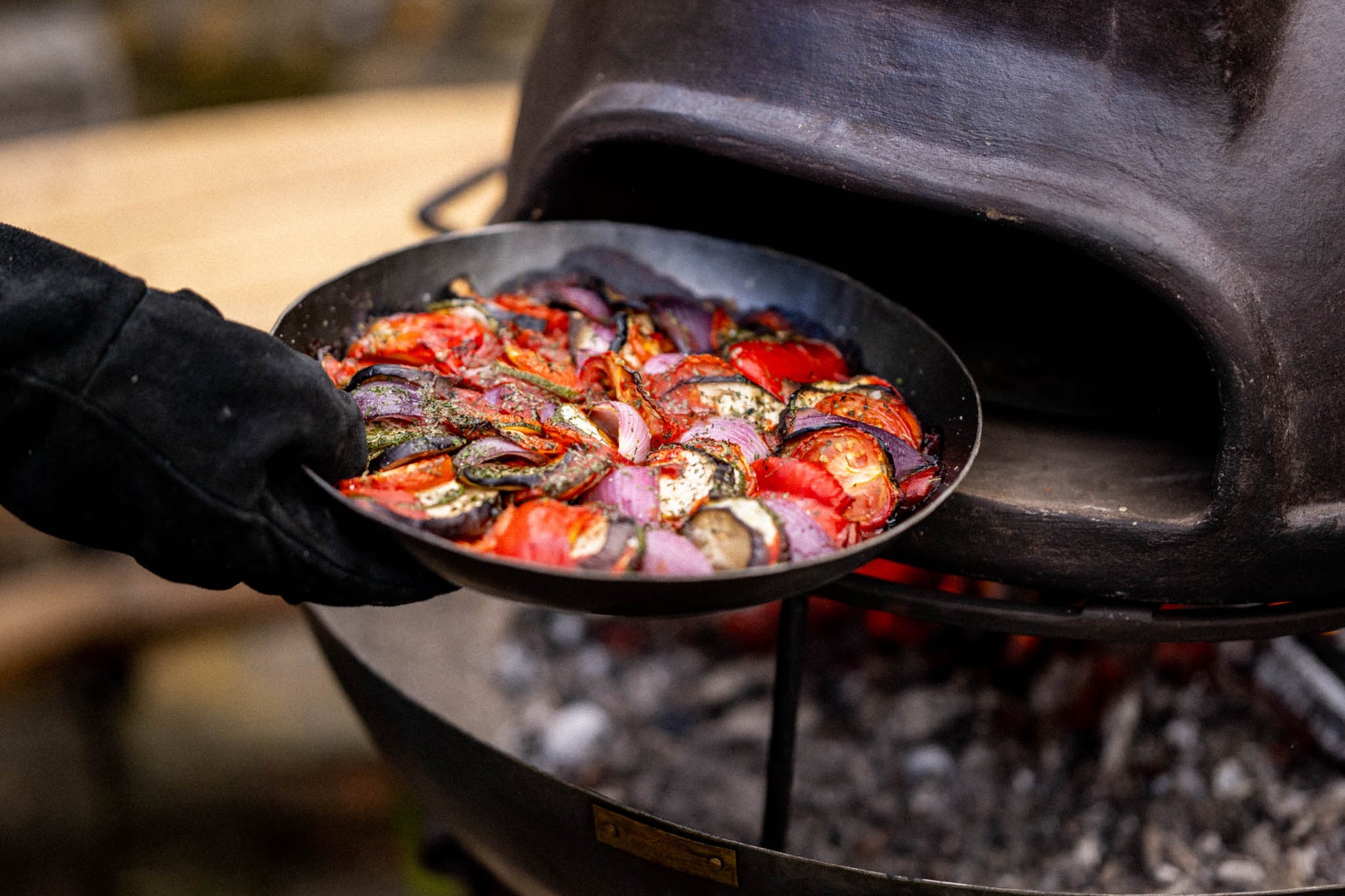 Close-up of a vegetable-filled oven dish being inserted into a rustic outdoor cooker with glowing embers underneath