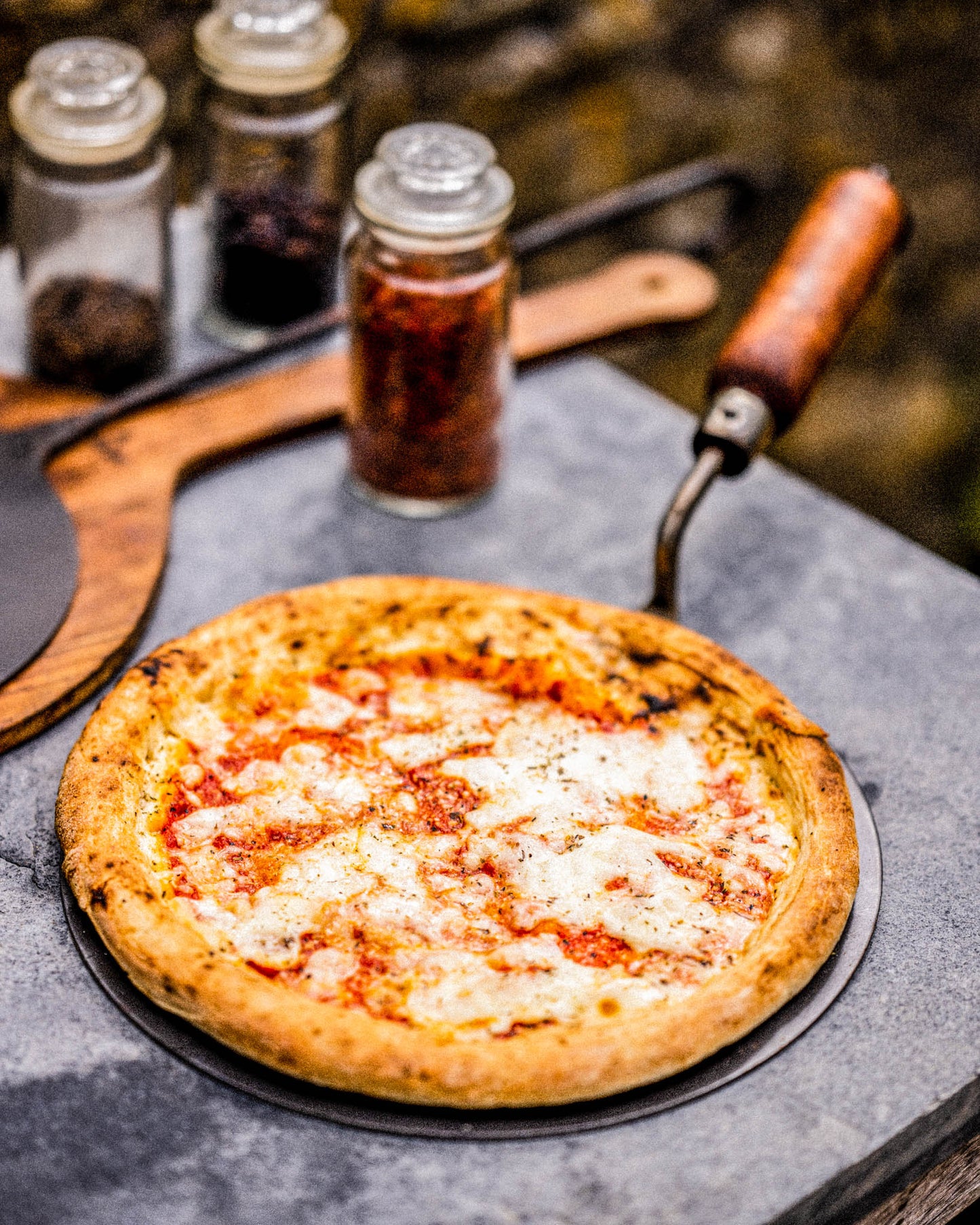 Pizza on a wooden-handled peel with melted cheese and tomato sauce, next to spice jars, on a pizza oven surface.