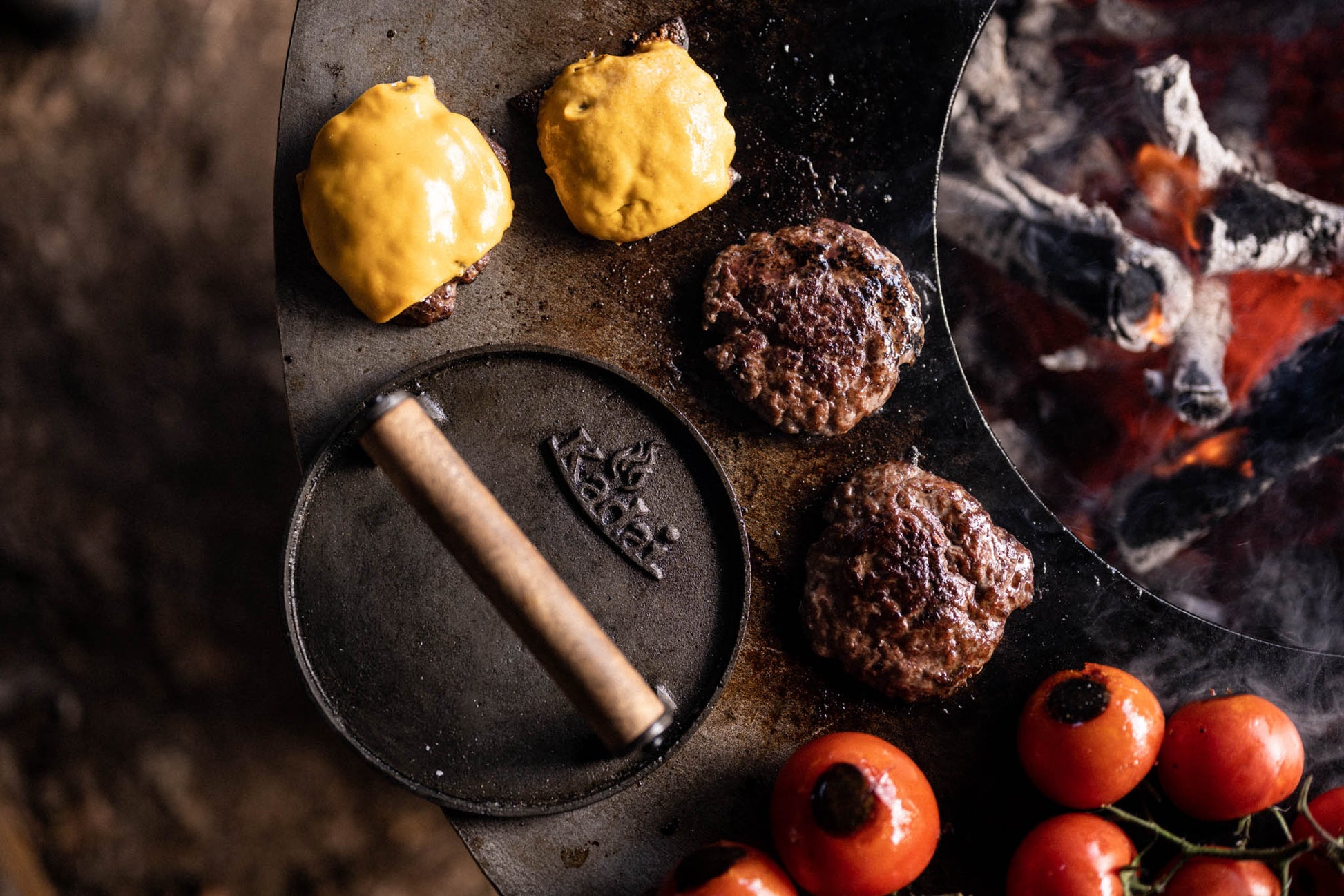 Smash burgers cooking on a plancha kadai with melted cheese, tomatoes and open fire