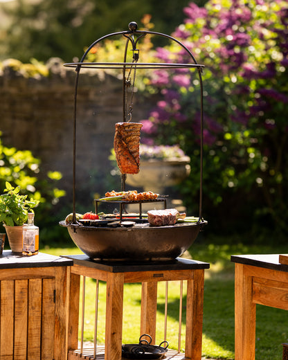 Modern outdoor BBQ plancha grill in a sunny garden setting, featuring hanging ribs suspended over an open fire with steaks, skewers, and vegetables cooking on a circular flat-top grill and raised grate