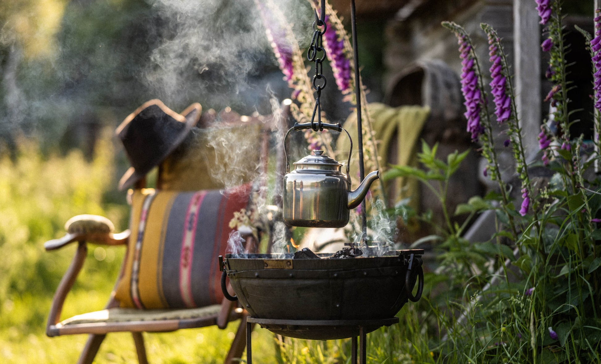 Portable BBQ hook holding a kettle over a fire pit in a garden setting with flowers and outdoor seating
