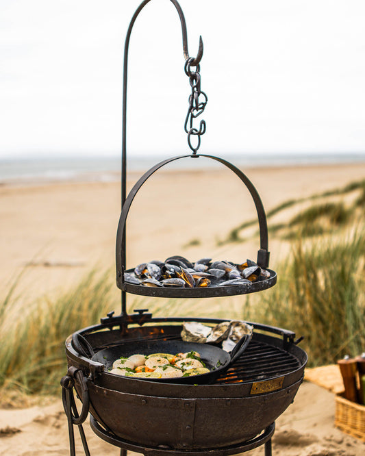 Portable BBQ skillet cooking scallops on a Kadai grill with hanging rack of mussels above on a beach