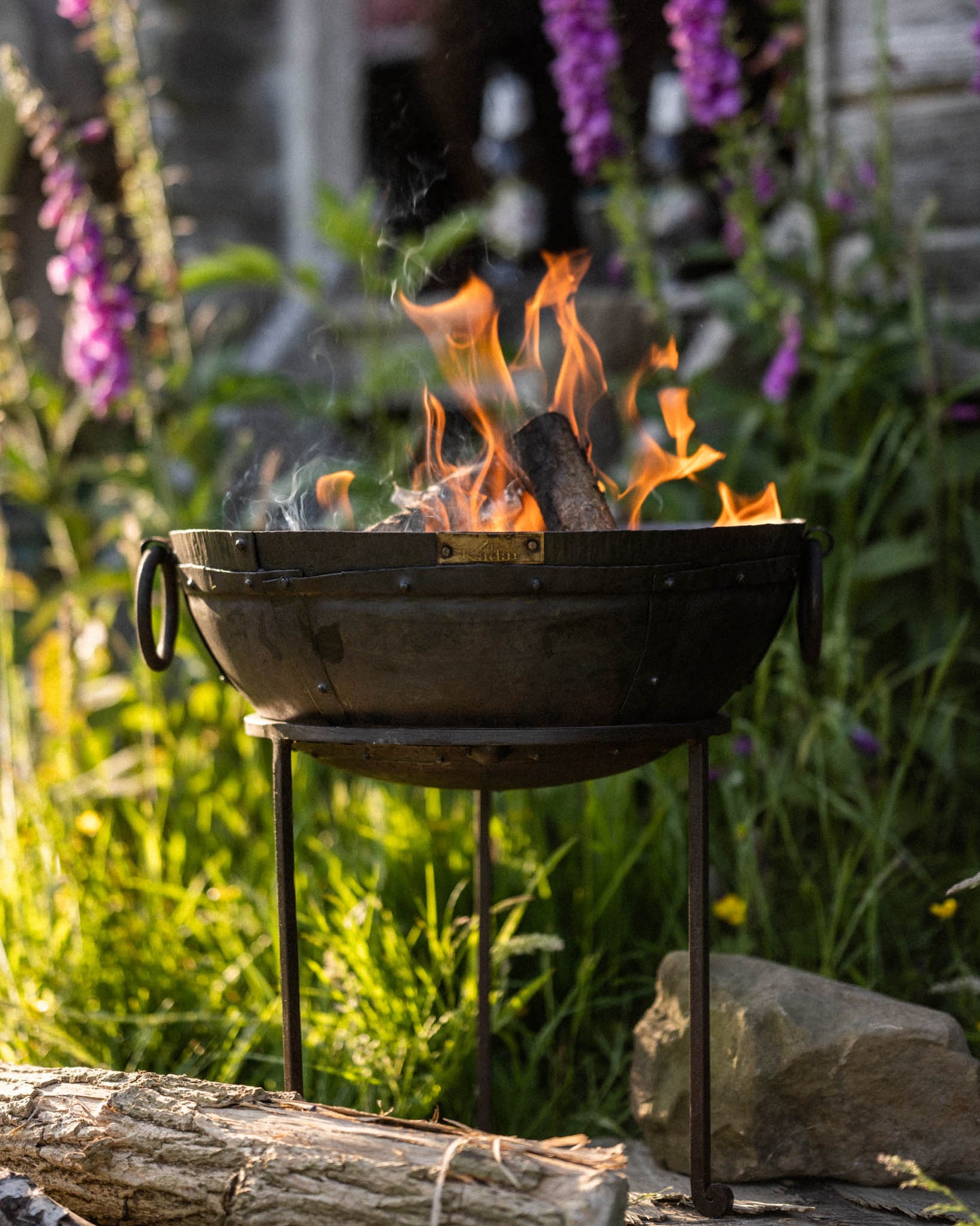 Portable BBQ fire pit with burning logs in a Kadai firebowl on a stand in a garden setting