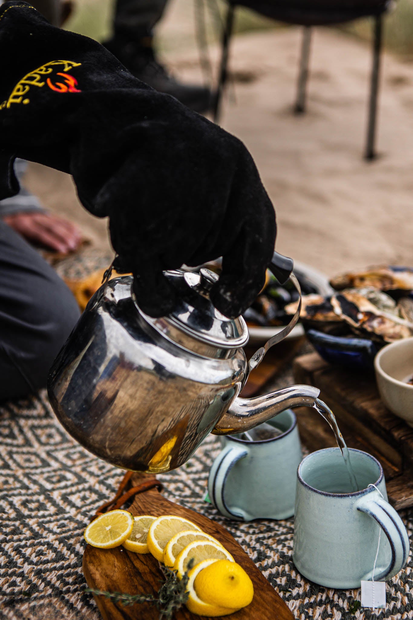 Camping BBQ kettle pouring hot water into mugs, with sliced lemon on a wooden board beside an outdoor setup