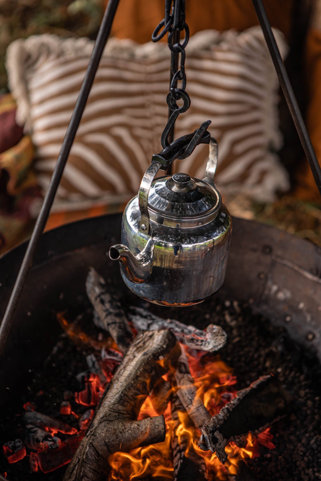 Camping BBQ kettle hanging over an open fire pit, suspended by a tripod chain above glowing embers