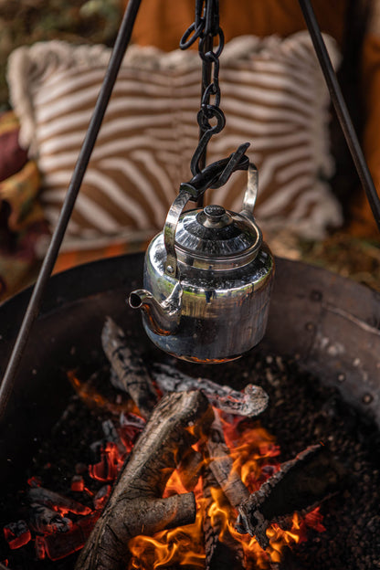 Camping BBQ kettle hanging over an open fire pit, suspended by a tripod chain above glowing embers