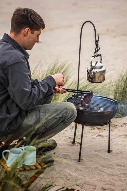 Foldable camping stool beside a portable BBQ on the beach with a man cooking over charcoal