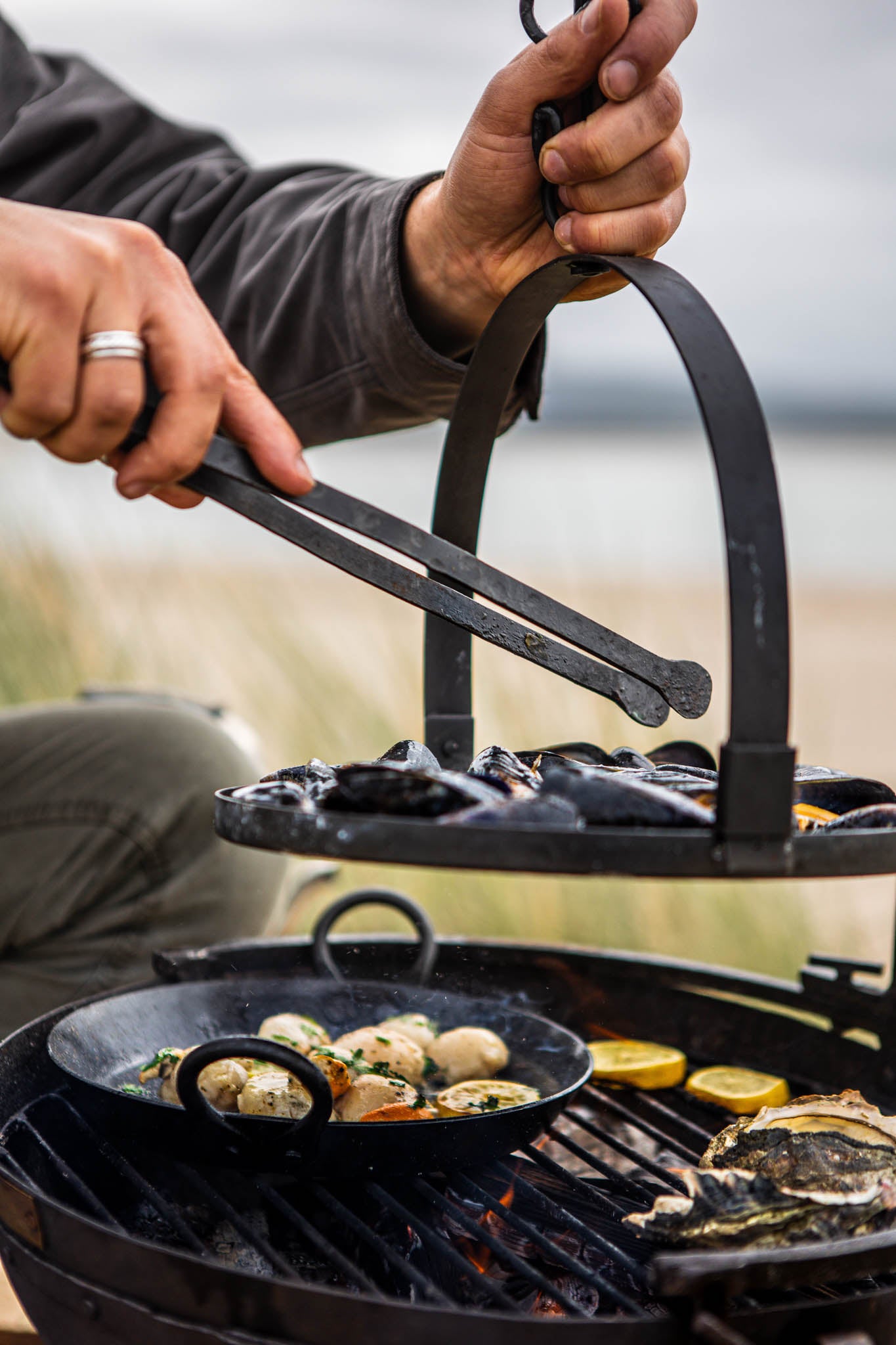 Portable BBQ skillet cooking scallops on a Kadai grill, with mussels on a hanging rack being lifted above the fire