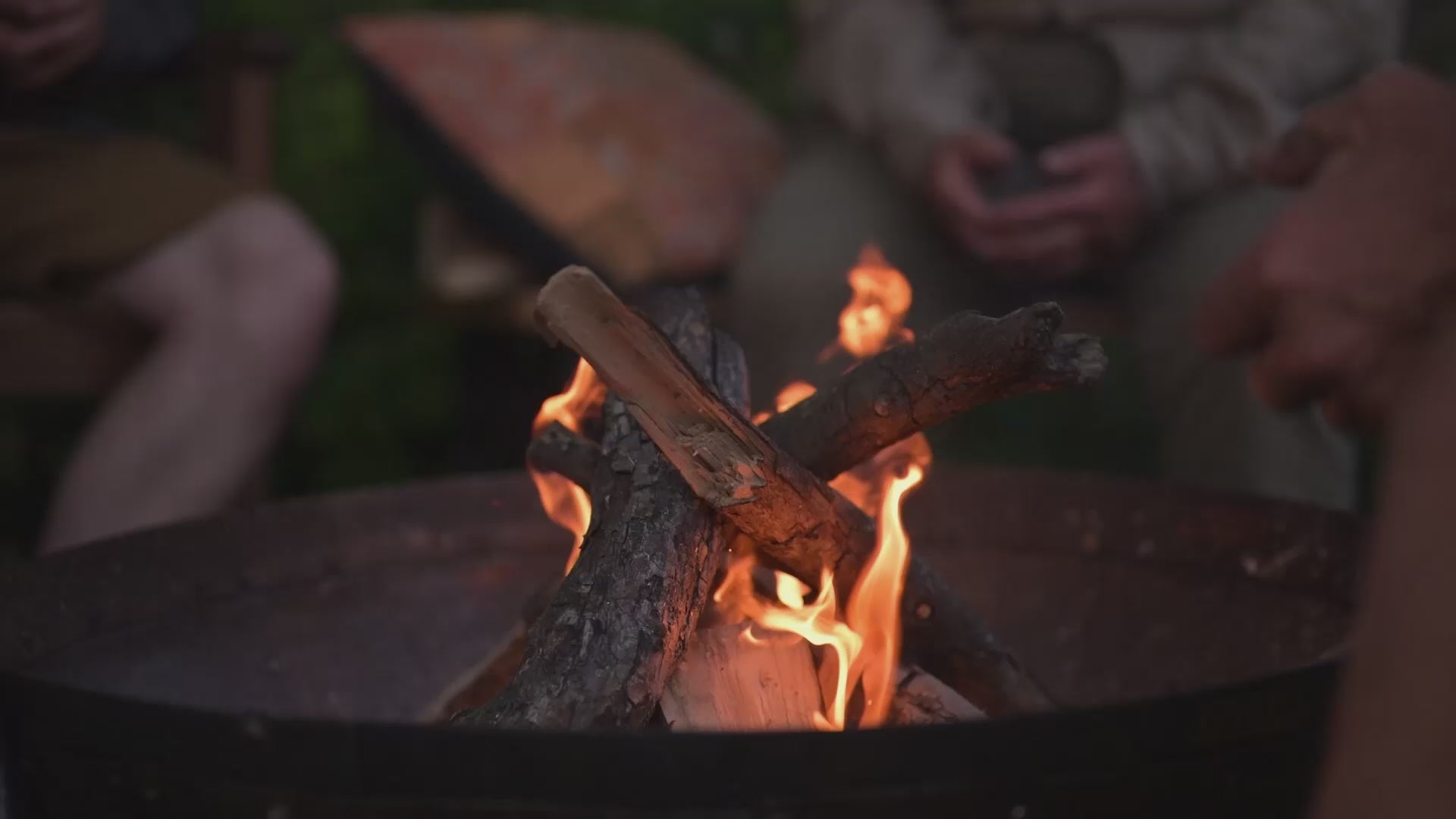 People gather around the warmth of flames with a fire inside a round metal fire pit, candles are lit and glasses of wine are in peoples hands.
