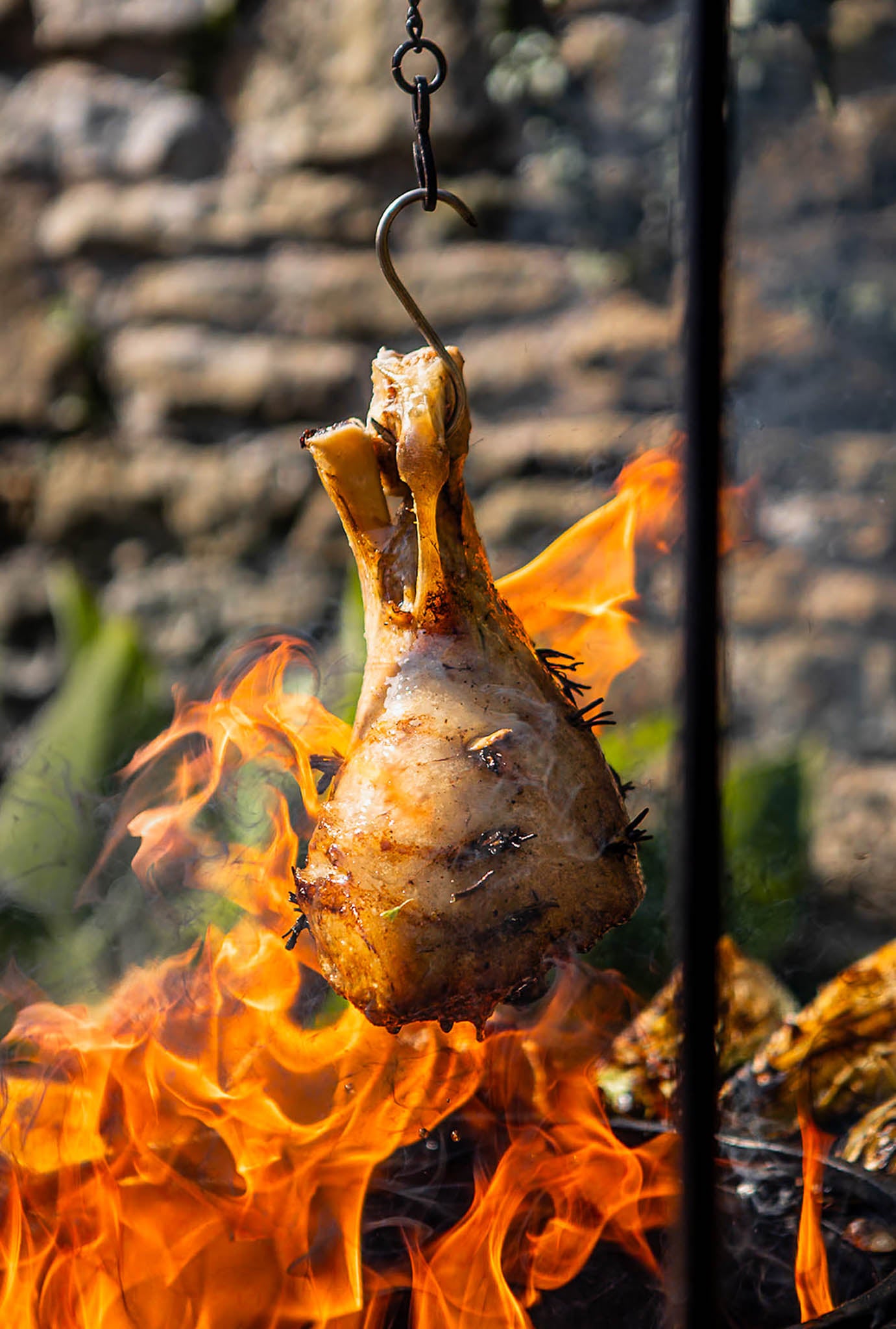 Roasted leg of lamb suspended from a tripod pulley hook over open flames on a BBQ plancha grill, with fire blazing beneath in an outdoor rustic cooking setting.