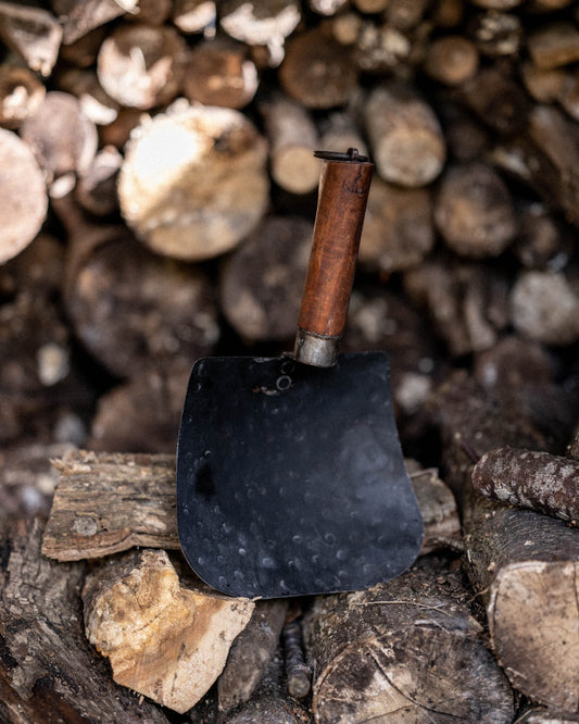 Black metal Kadai shovel with a wooden handle resting on stacked firewood logs outdoors.