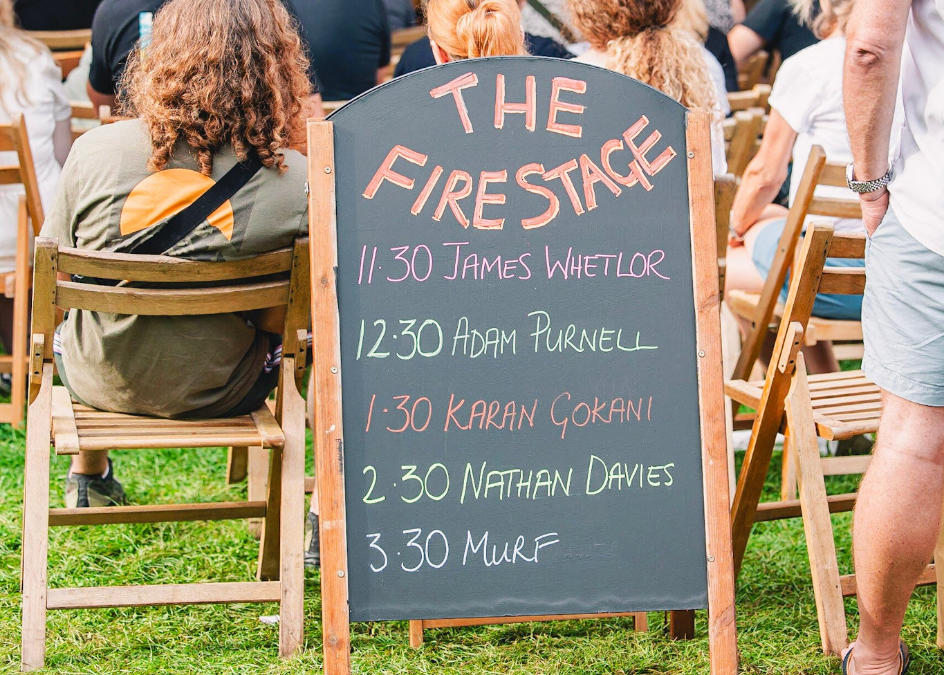 Outdoor event with a chalkboard sign titled 'The Fire Stage' listing names and times.