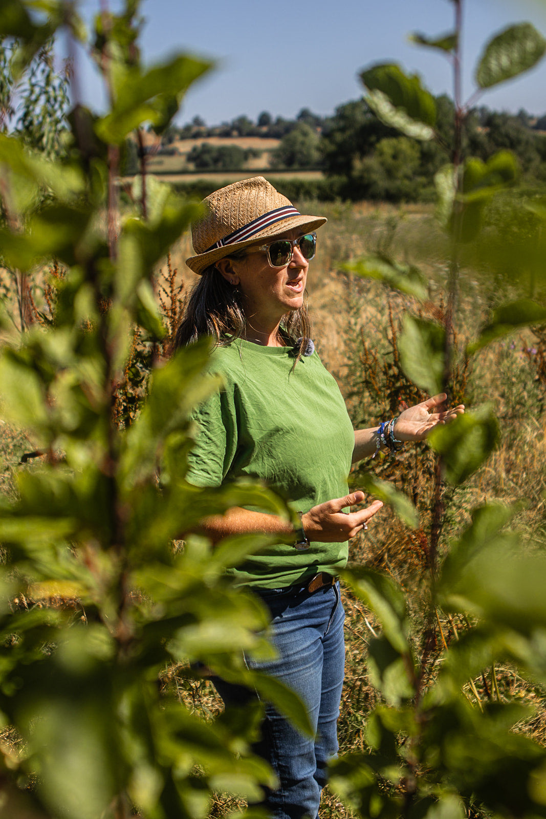 A woman wearing a straw hat, sunglasses, a green t-shirt, and blue jeans stands in a sunny field of leafy plants, gesturing with her hand toward the vegetation.