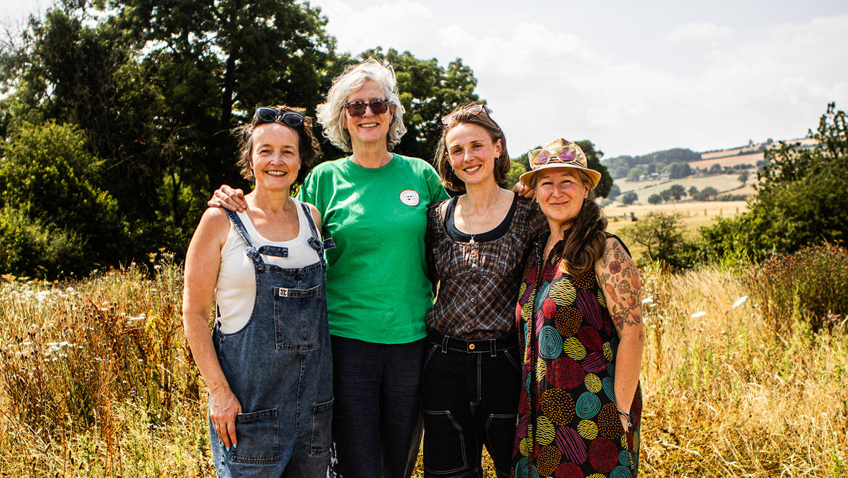 Four women who founded shropshire good food partnership standing together in a grassy outdoor field with trees and hills in the background, wearing casual clothing including denim overalls, a bright green T-shirt, a patterned dress, and a colorful hat.