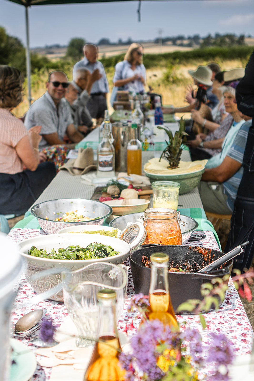 A long outdoor dining table set with bowls of food, bottles, glasses, and floral tablecloths, surrounded by a group of people seated and standing in a field under a canopy. Shropshire Good Food Trail launch day.