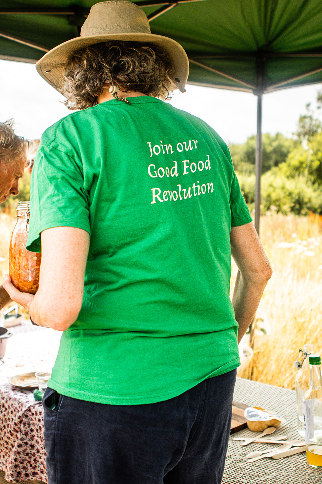 A person wearing a bright green T-shirt with the white text 'Join our Good Food Revolution' on the back, standing outdoors under a green canopy at a table.