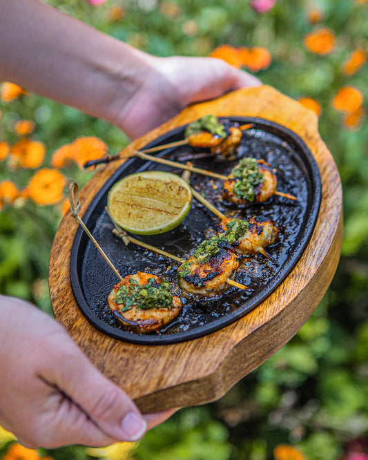 Hands holding a wooden sizzler plate with grilled shrimp skewers, chimichurri sauce, and a grilled lime, outdoors with flowers in the background.