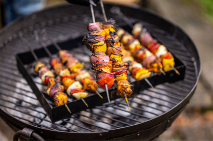Grilled vegetable and meat skewers being lifted from a BBQ skewer rack over hot charcoal on an outdoor grill