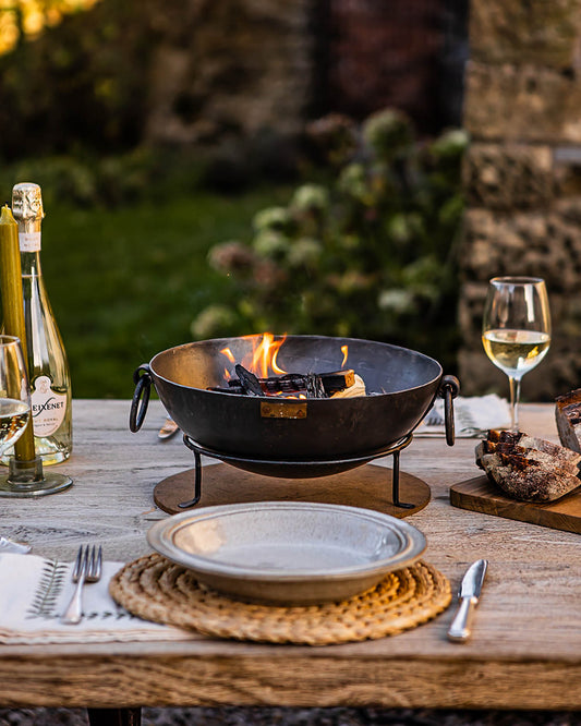 Outdoor BBQ table with Kadai fire bowl burning at centre, surrounded by plates, wine and rustic dining setup