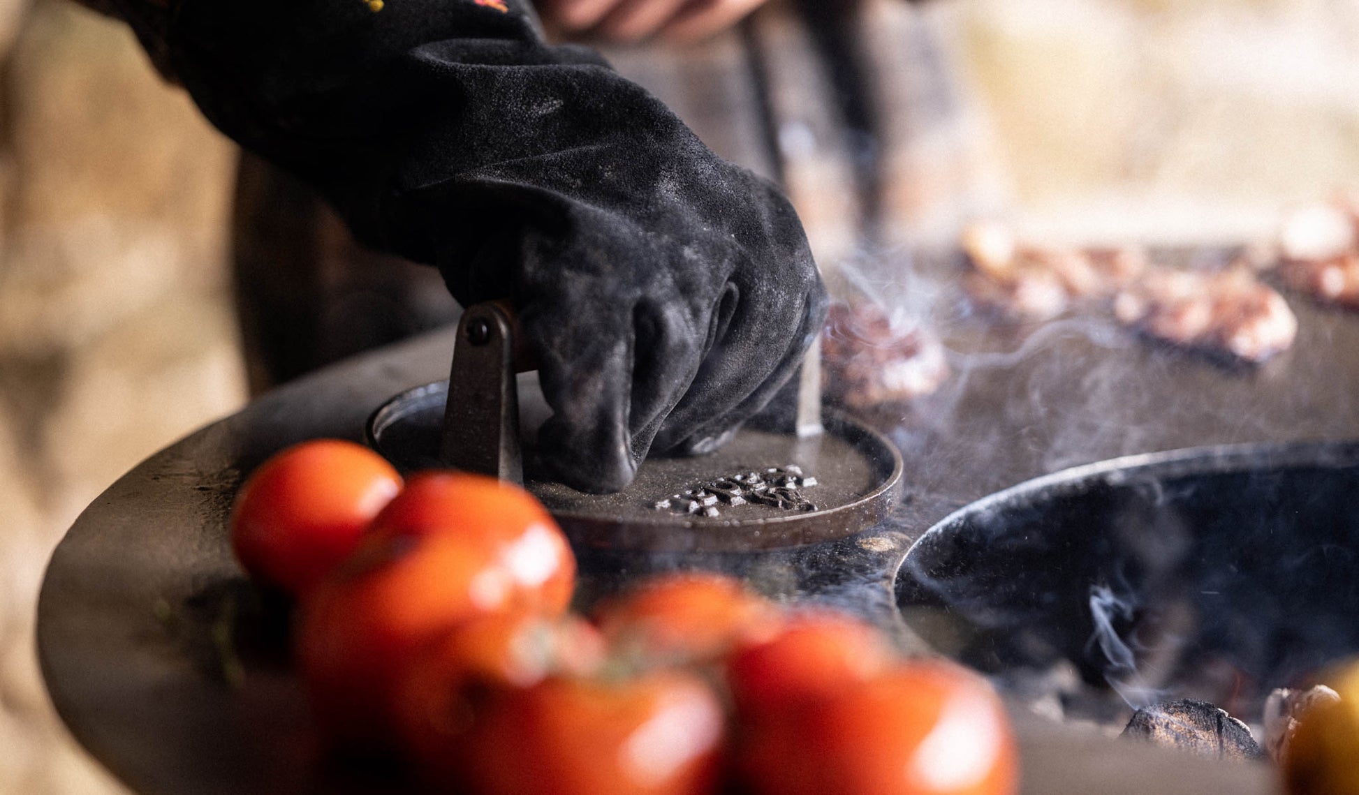 Gloved hand pressing a burger with a burger press on a hot grill, steam rising, tomatoes in the foreground.