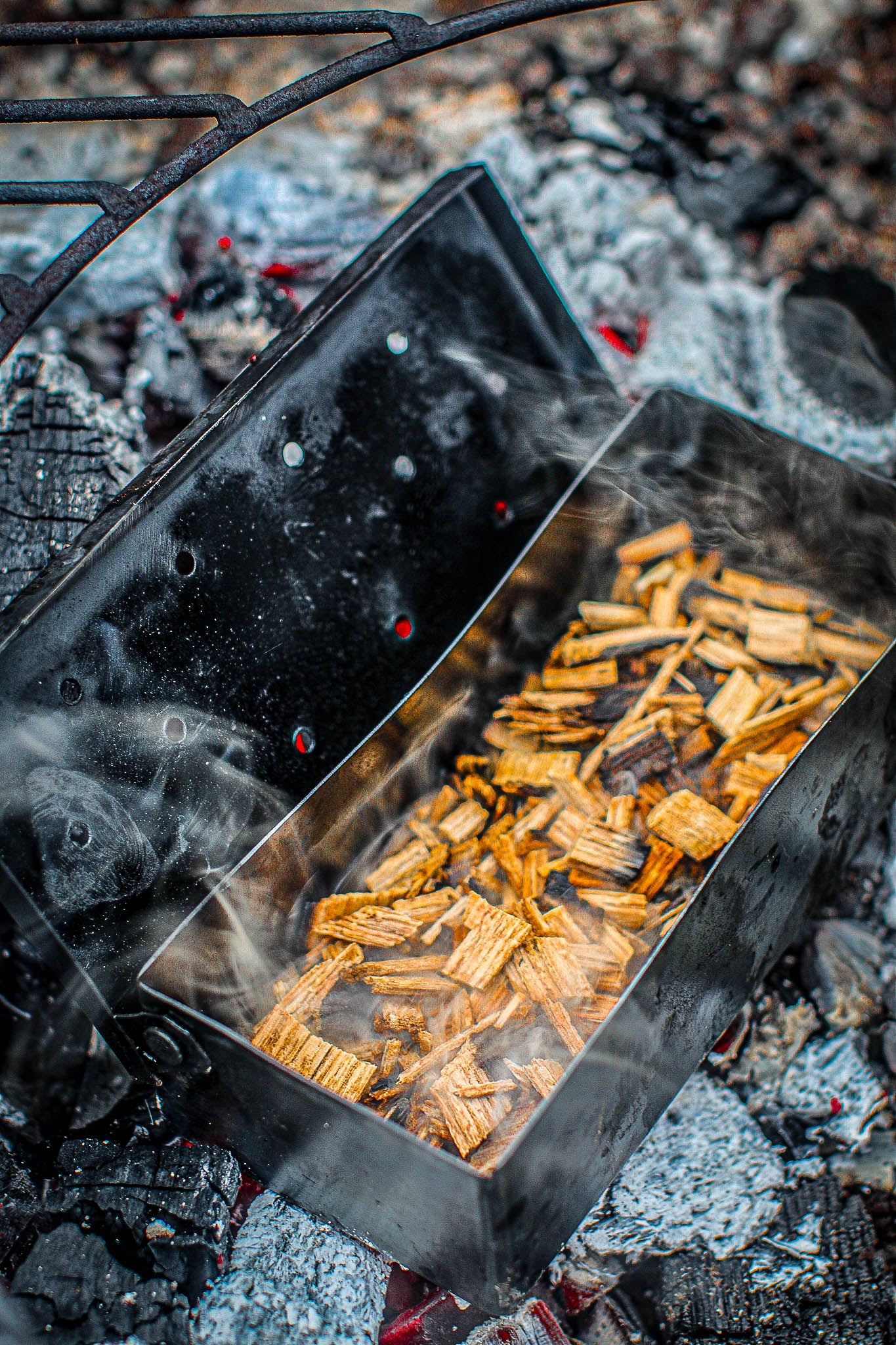 Smoker box filled with wood chips, releasing smoke over hot coals on a grill.