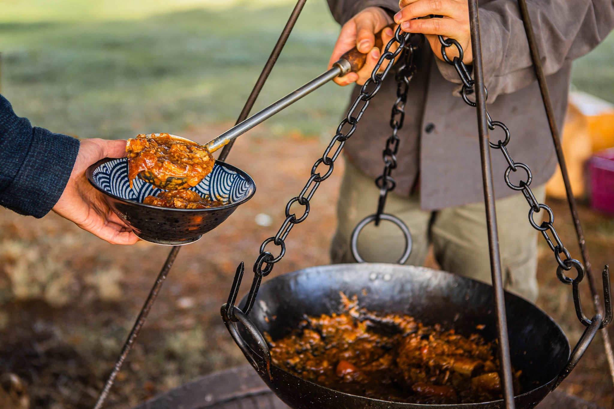 Stainless steel barbecue ladle with wooden handle being used to dish up hearty fire-cooked food at an outdoor gathering