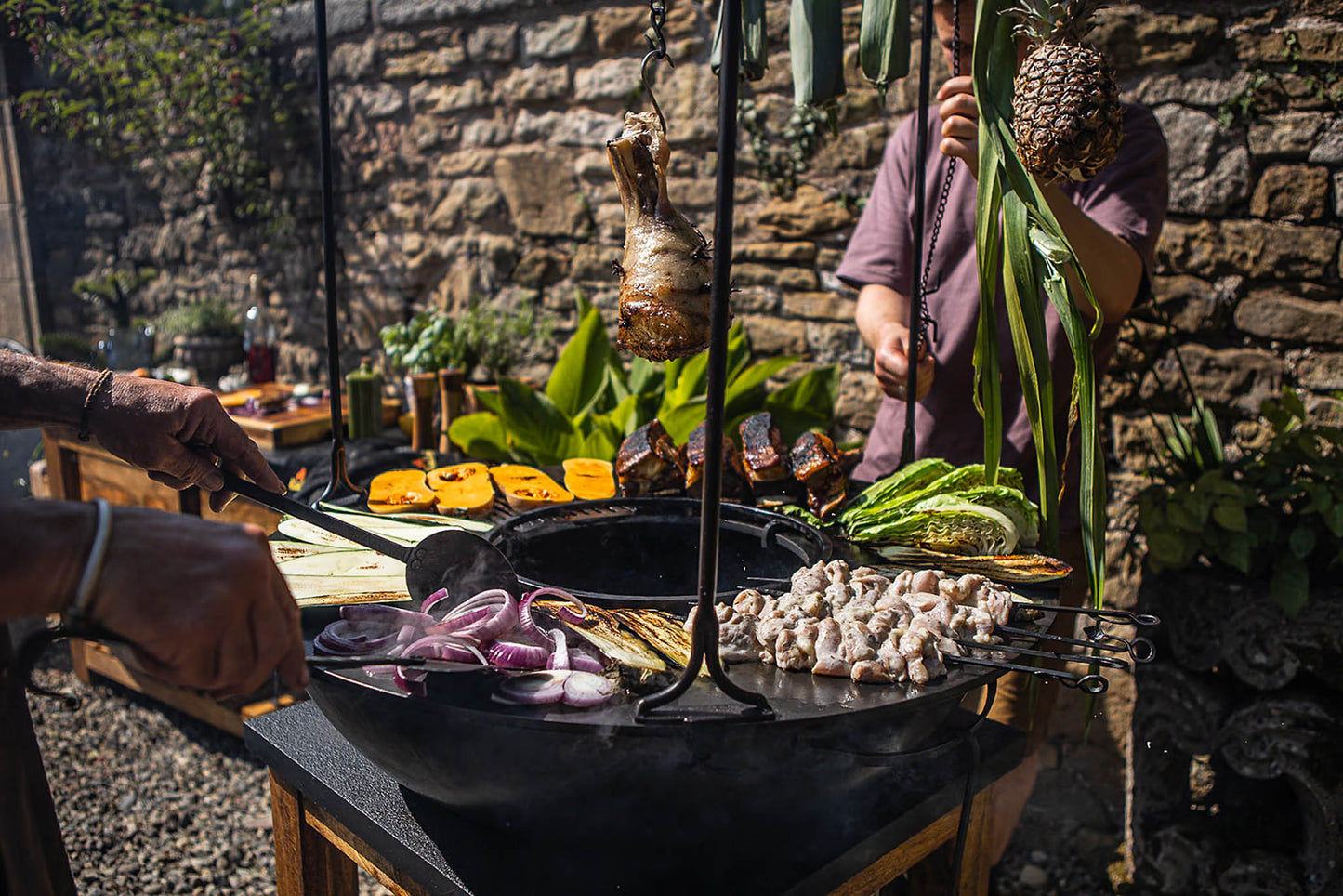 Outdoor plancha kadai grill cooking vegetables, skewers and hanging meat over open fire